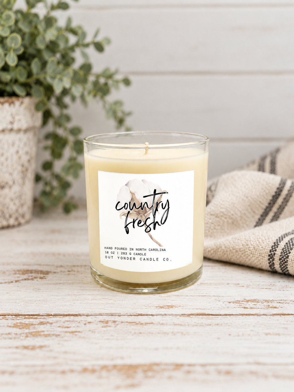 A single-wick soy candle in a clear glass jar with a cream-colored wax and a white label that reads 'country fresh' with a floral illustration. The candle is styled on a rustic white wooden surface next to a beige striped cloth and a potted green plant.